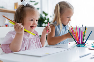 happy toddler kid with down syndrome holding pencils near blurred blonde girl