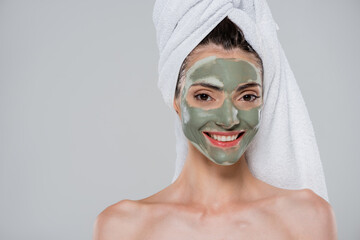 joyful young woman with towel on head and green clay mask on face isolated on grey
