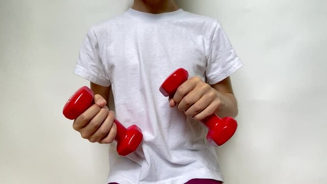 A girl in a white T-shirt slowly moves two red dumbbells weighing 1 kg 