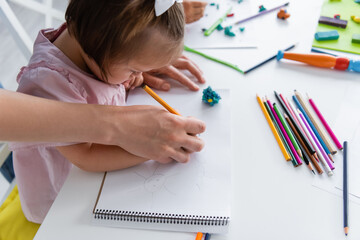 teacher assisting disabled girl with down syndrome drawing in private kindergarten