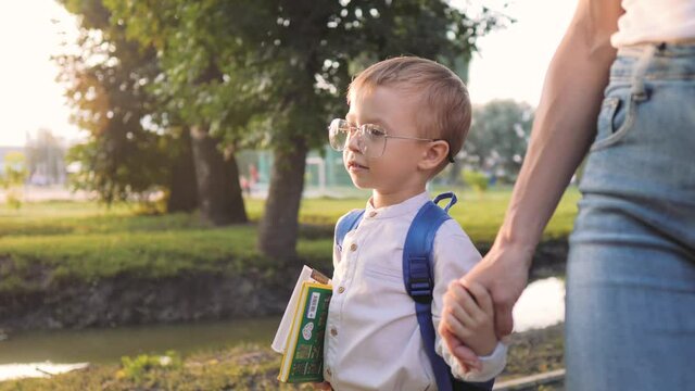 First time to school. Happy cute clever boy in glasses with school bag and book in his hand. Back to school. Parent and pupil of primary school go hand in hand. Mother supports her son before study.
