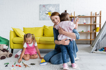 happy kindergarten teacher hugging toddler girl with down syndrome near preschooler child playing with toys in playroom