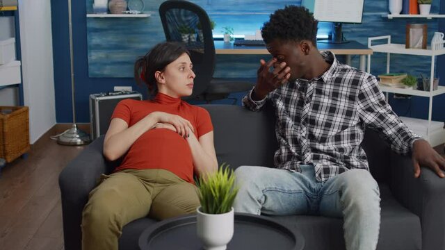 Interracial couple talking about child and parenthood while sitting on couch at home. Mixed race partners expecting baby feeling cheerful. Caucasian pregnant woman and black husband
