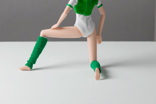 A faceless doll in a tracksuit and green leggings performs kneeling exercises against a gray wall. Anti-cellulite concept, sports activities.