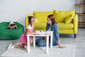 smiling kindergarten teacher looking at girl near papers and color pencils