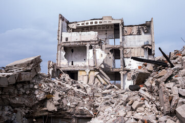 A destroyed building against a blue sky with gray clouds with a pile of concrete rubble in the...