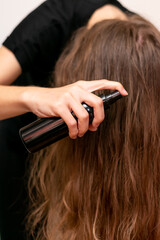 Young woman hand holds bottle and spraying her wavy blond long hair. Hair care for making it shiny,...