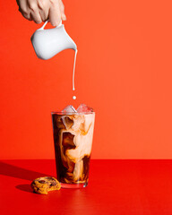Iced coffee on a bright red background with a hand pouring cream into the coffee