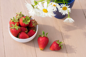 White bowl full of strawberries and white flowers of chamomile with long shadows on bright sunlight