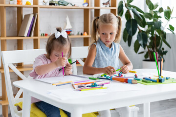 blonde girl looking at disabled toddler child with down syndrome drawing in private kindergarten