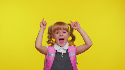 Attractive little teenage girl kid in school uniform wears pink backpack showing thumbs up and pointing up at copy space for promotional content on yellow studio background. Education, back to school