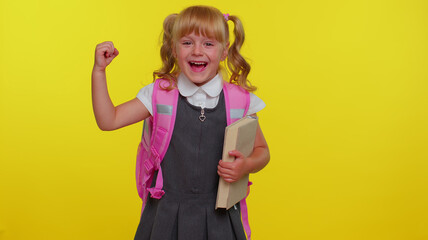 Happy little blond teenage student girl kid in school uniform celebrate success win scream rejoices doing winner hands gesture say Yes. Yellow studio wall background. Education, back to school, study