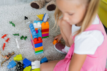 blurred girl in pink dress playing building blocks on carpet in kindergarten playroom