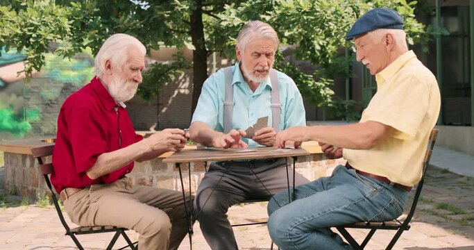 Full length view of the three caucasian senior men best friends on retirement playing cards in the yard at the table. Outside. Leisure games and retirement concept