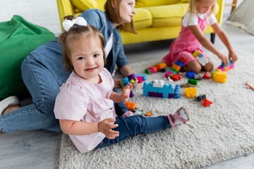 toddler kid with down syndrome looking at camera while playing with blurred girl and kindergarten teacher on carpet