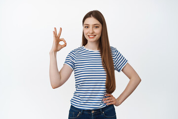 Very good. Satisfied girl smiles, shows okay, OK sign, stands against white background