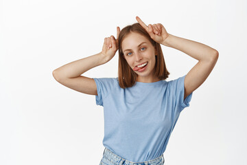 Young carefree blond woman show tongue, bull devil horns gesture on head, smiling carefree, having...