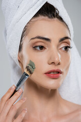 young woman with towel on head applying clay mask with cosmetic brush isolated on grey
