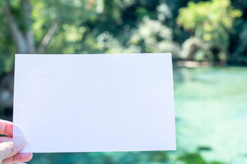 Closeup of woman's hand holding blank paper greeting card. Invitation mockup in sunlight. Blurred background with crystal clear water and lush greenery. Selective focus.Summer party concept