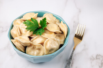 Blue deep bowl with meat dumplings with herbs on a white marble table with a gold fork. Simple table setting, light background.