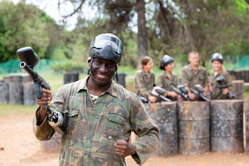 Fototapeta premium Smiling afro american man paintball player in camouflage posing with gun outdoors