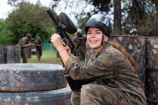Young smiling woman paintball player in camouflage posing with gun outdoors