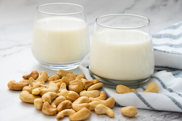 Glasses of vegan milk with heap of cashew nuts on kitchen towel on marble background. Lactose free non-dairy milk drink. Alternative types of milks. Vegan food and healthy eating concept.