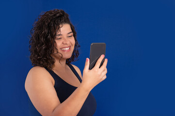 brazilian young woman with curly hair smiling while looking to a smartphone on a blue background with copy space