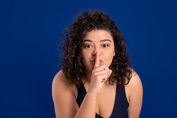 young Woman on blue background showing a sign of silence. Shut Your Mouth. Finger on Lips. Shut up.