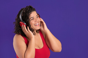happy young woman with curly hair and red shirt listening music with headphones on a purple background with copy space
