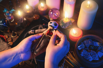 Sorceress or witch sticks needles into voodoo doll at ritual table with pentagram, burning candles and other occult objects, top view. Voodoo witchcraft, spirituality and occultism concept. - obrazy, fototapety, plakaty