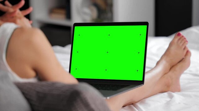 Back view of mature lady lying on bed and using modern laptop with green chroma key screen. Concept of people, leisure time and technology.
