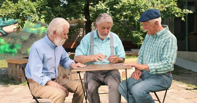 Waist up portrait view of the old caucasian man choosing a card for his turn while his friends making the move. Outdoor.