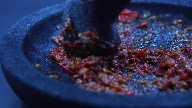 The hand is grinding the red chili sauce in a stone mortar. Chef preparing Indonesian traditional spicy hot chili sauce