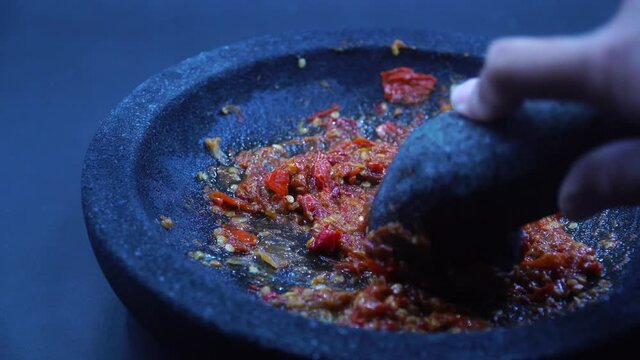 The hand is grinding the red chili sauce in a stone mortar. Chef preparing Indonesian traditional spicy hot chili sauce. Zoom in