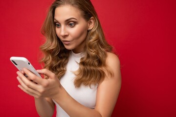 Closeup portrait of serious concentrated beautiful young blonde woman wearing white t-shirt isolated over red background using smartphone and texting message via mobile phone looking at gadjet screen