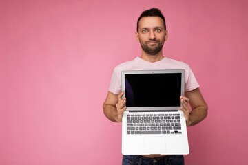 Handsome surprised man holding laptop computer looking at camera in t-shirt on isolated pink background