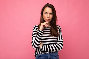Shot of young serious thoughtful nice cute brunette woman with sincere emotions wearing casual striped sweater isolated on pink background with empty space