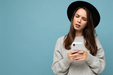 Beautiful young asking dissatisfied brunette woman wearing black hat and grey sweater holding...