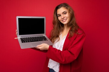 Photo of Beautiful smiling happy young brunette woman holding computer laptop with empty monitor screen wearing red cardigan and white blouse looking at camera isolated over red background