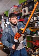 Adult glad cheerful positive male is standing with new electric brush cutter in tools store