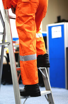 A Worker Is Climbing And Holding A Ladder Wearing Safety Shoes And An Orange Coverall. He's Fixing Something In The Office