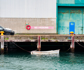 Old dinghy tied to wharf with old shed background lifebuoy on wall and no parking sign with two cars