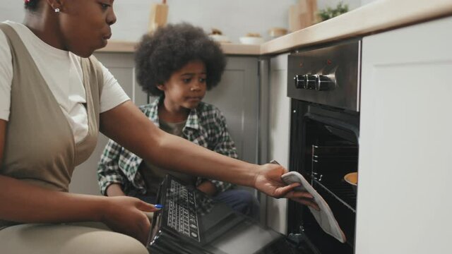 Slowmo shot of young African-American woman and her cute little son taking delicious homemade apple pie out of oven enjoying its taste