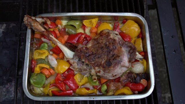 Chef cooking a delicious leg of lamb on barbecue with colors peppers and potatoes in a metal tray. Closeup of tasty goat meat in BBQ of garden home at holiday vacations. Roast grill food in Spain.-Dan