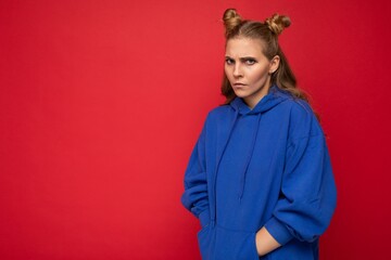 Portrait of touchy sad young beautiful winsome blonde woman with two horns with sincere emotions wearing stylish bright blue hoodie isolated on red background with copy space