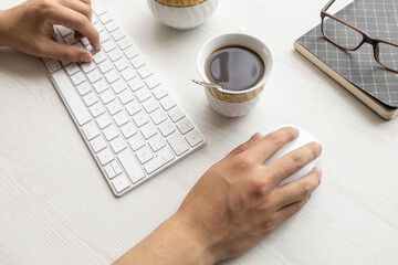 on a desk the hands of a person typing on a modern keyboard and using his mouse, around him glasses, cup of coffee, lifestyle at work