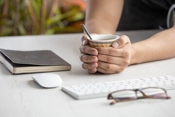 hands of a person holding a cup with hot coffee on a wooden desk, around a notebook, glasses, computer mouse, and keyboard. Office items