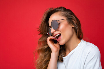 Closeup shot of charming happy young dark blonde curly woman isolated over red background wall wearing casual white t-shirt and stylish sunglasses looking to the side