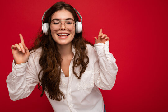 Closeup photo of attractive positive smiling young brunet woman wearing white shirt and optical glasses isolated over red background wearing white wireless bluetooth headsets listening to music and